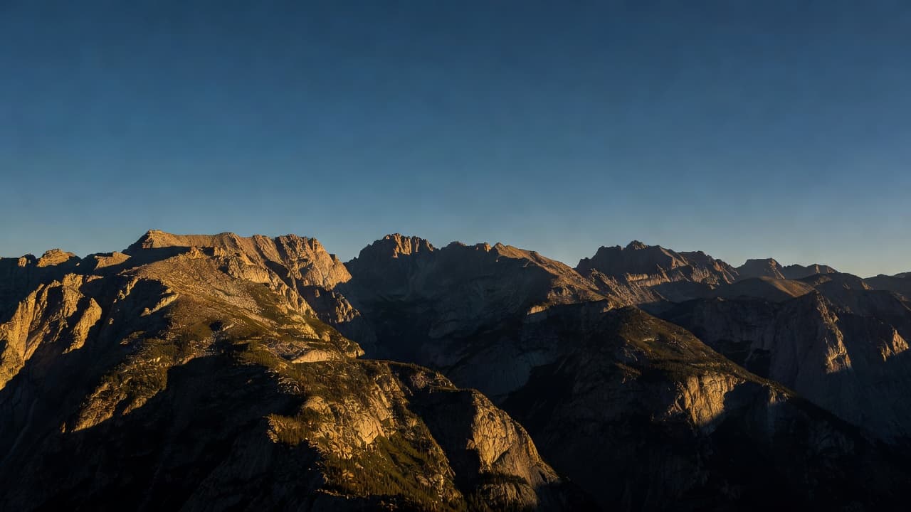 Sharp rocky peaks rise against a vast, clear sky during golden hour. Deep, elongated shadows cast dramatic textures across the expansive valley floor, emphasizing the immense scale of the landscape.